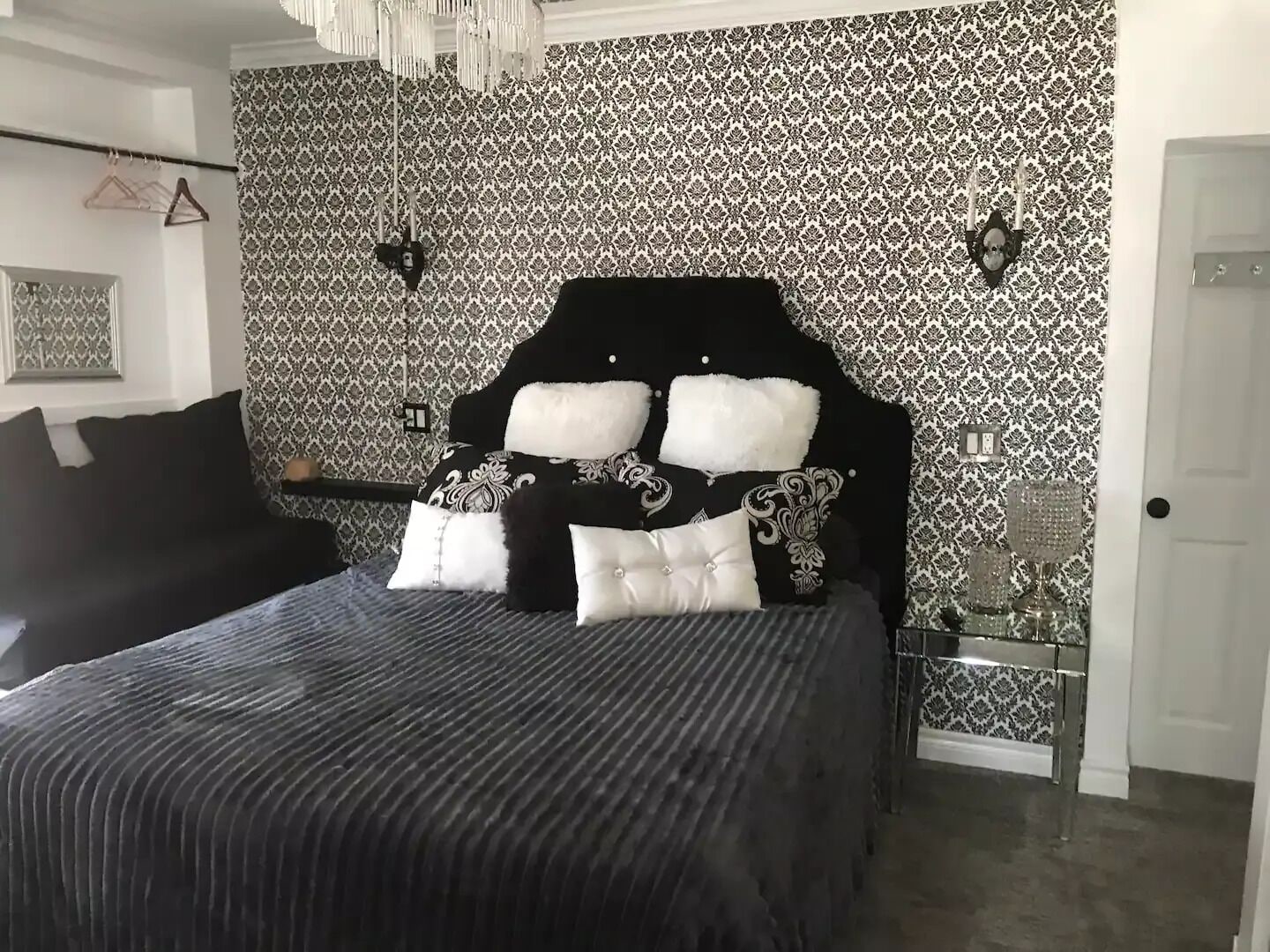 A bedroom with a black and white patterned wallpaper, a black headboard with white cushions, a black sofa, and a bedside table with a lamp.