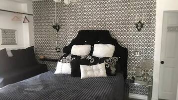 A bedroom with a black and white patterned wallpaper, a black headboard with white cushions, a black sofa, and a bedside table with a lamp.