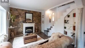A living room with a stone fireplace, a white staircase, and a bookshelf. - Private house/large hottub/sauna & optional "Adult/Red room" (Rothesay)