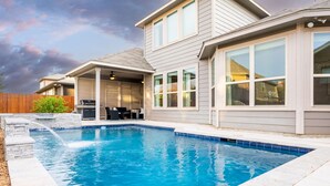 Ein Swimmingpool mit Steinbord, eine überdachte Terrasse mit Gartenmöbeln und ein Haus mit großen Fenstern und einer grauen Fassade.