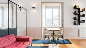 En moderne stue med en rød sofa, et rundt træbord med barstole, et stort vindue og en sort vægmonteret hylde med planter.