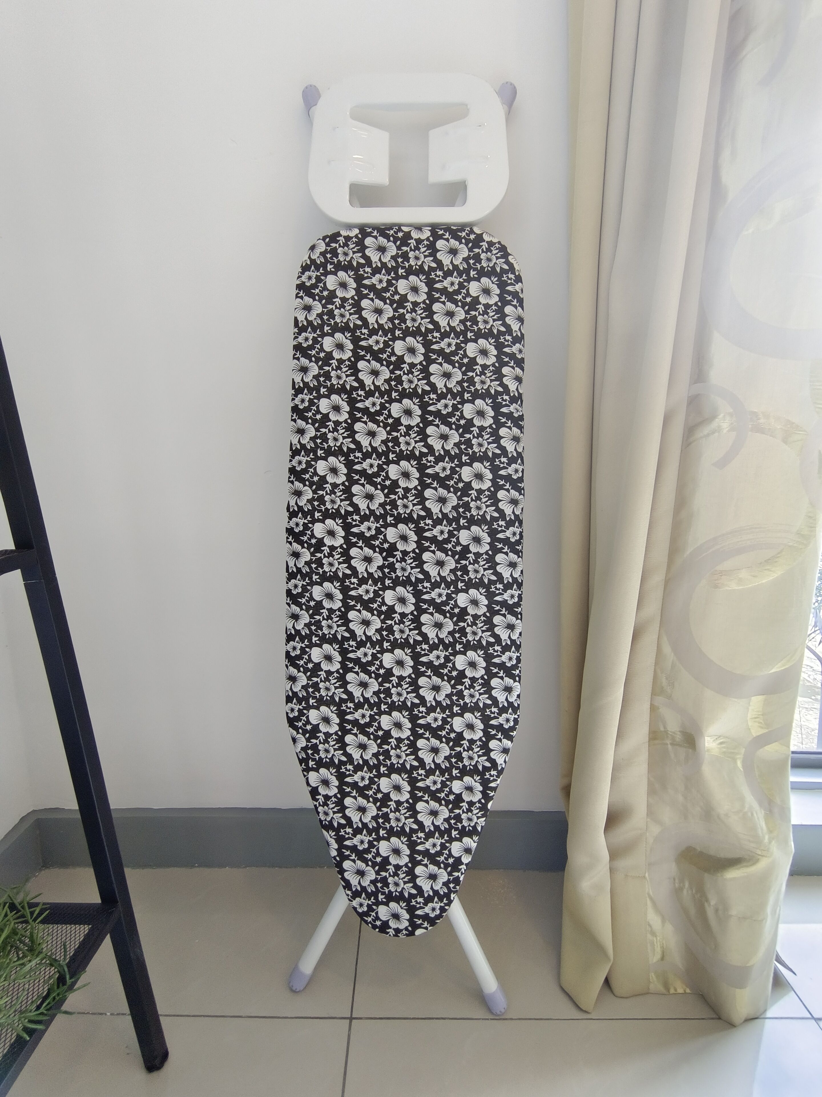 Una tabla de planchar plegable con estampado floral, ubicada frente a una pared con ventana y cortinas.