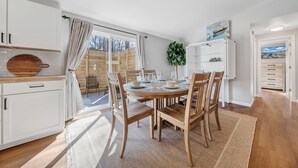 Een moderne keuken met witte kastjes, een houten eettafel en stoelen. Er is een groot raam met gordijnen, een plant en uitzicht op buiten.