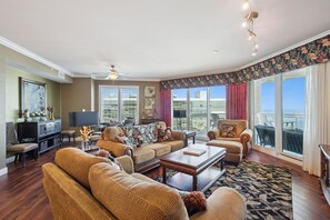 Un salón amplio con un gran sofá, una mesa de centro y un televisor. Grandes ventanales ofrecen vistas al exterior.