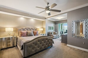 Une chambre à coucher avec un lit doté d’une tête de lit capitonnée, deux tables de chevet avec des lampes, un ventilateur de plafond et un grand miroir au mur.