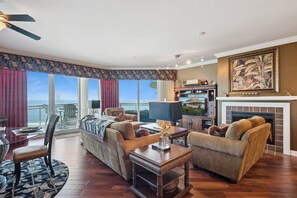 A spacious living room with a large window overlooking the ocean, a fireplace, and a television. - Prestigious Luxury Coastal Oceanview & Sunset Condo (Daytona Beach)
