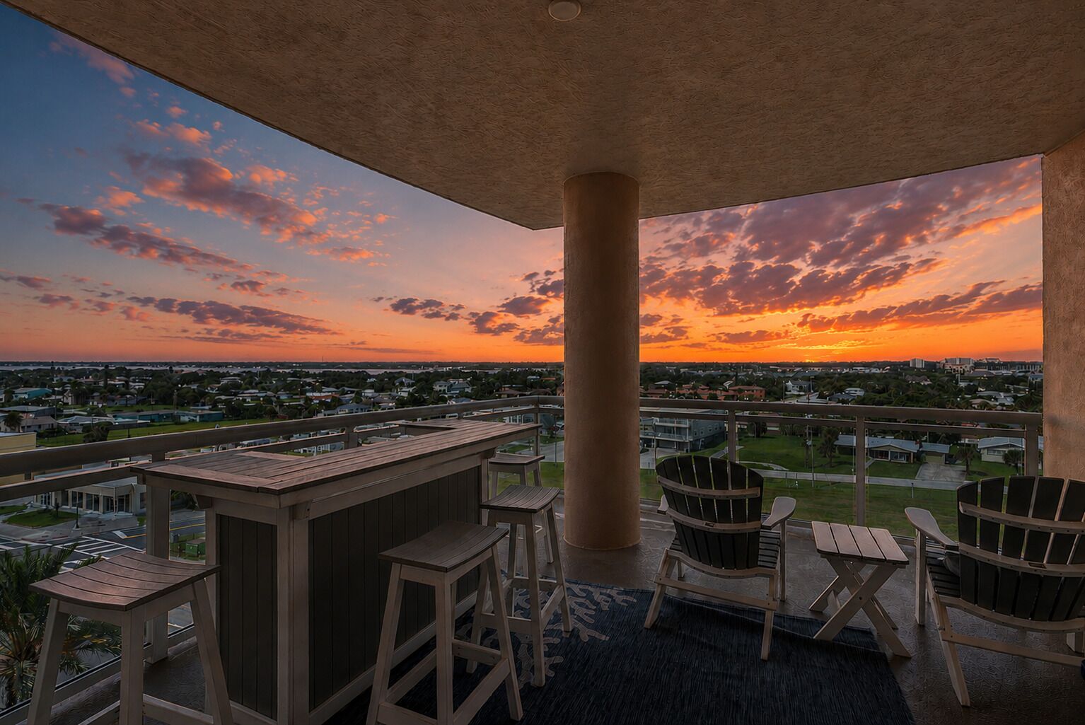An outdoor balcony with a view of a sunset over a residential area, featuring a wooden table and chairs.