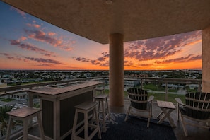 Un balcón al aire libre con vistas a una puesta de sol sobre una zona residencial, que incluye una mesa y sillas de madera.