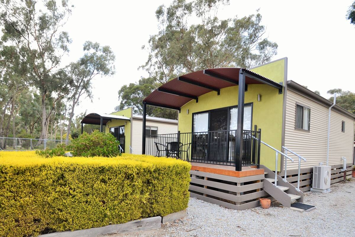 A caravan-style accommodation with a covered outdoor seating area, a gravel driveway, and a well-maintained hedge.