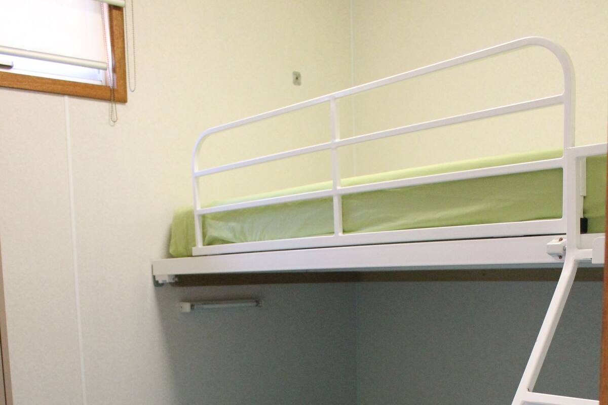 A bunk bed with a green mattress and a blue mattress, wooden flooring, and a window with blinds.