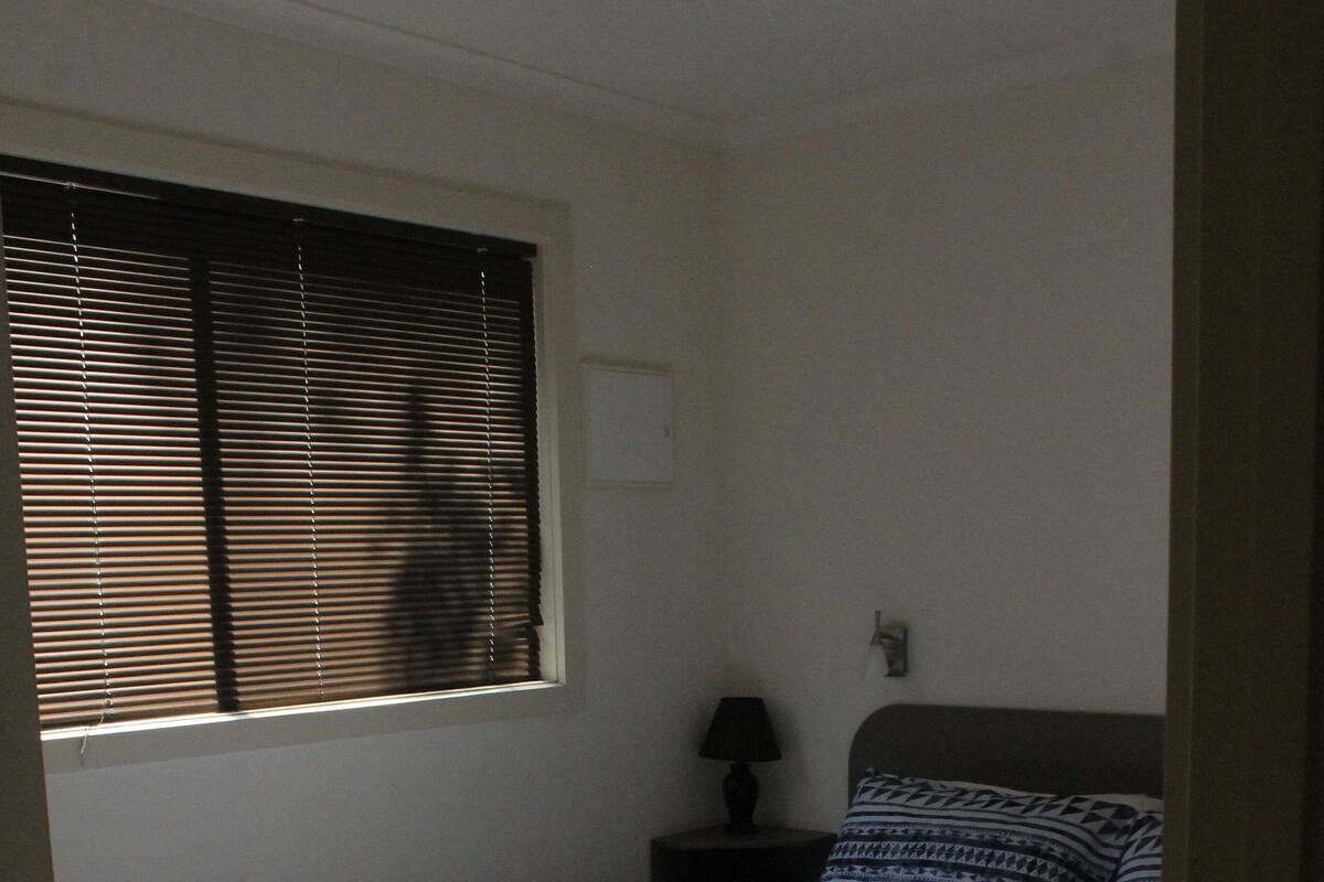 A bedroom with a bed, a window with blinds, a bedside table with a lamp, and a wall-mounted light fixture.