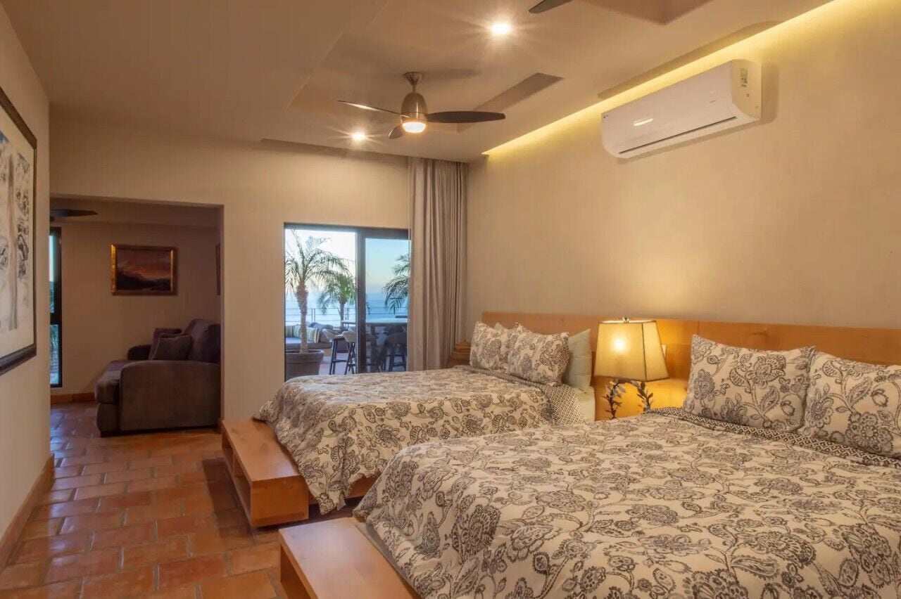 A hotel room with two beds, a ceiling fan, an air conditioner, a balcony with a view, and a dining area.