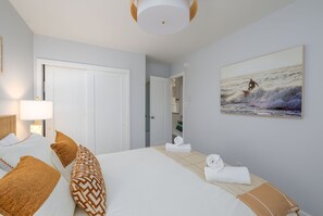 A hotel room with a bed, a painting of a surfer, a door, and a lamp.