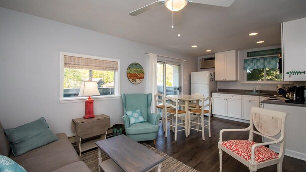 A living room with a sofa, a coffee table, a teal armchair, and a dining table with chairs. There is a kitchen area with white cabinets and a window with a view of the outdoors. - Park and Walk! Downtown Rehoboth Beach Condo (Rehoboth Beach)