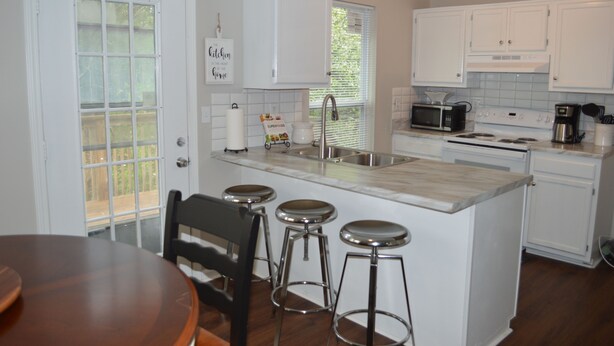 A kitchen with white cabinets, a microwave, a stove, and a sink. There are three bar stools and a wooden table. - Spacious Home Near Atlanta, Sweetwater Park, Six Flags and Airport (Lithia Springs)