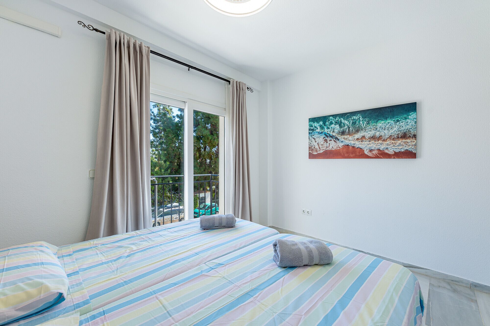 A bedroom with a large window, a bed with striped bedding, and a painting of waves on the wall.
