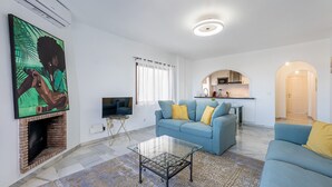 Ein modernes Wohnzimmer mit einem blauen Sofa, einem Glastisch und einem Flachbildfernseher. Durch einen Bogen ist eine Küche zu sehen, und an der Wand hängt ein Gemälde.