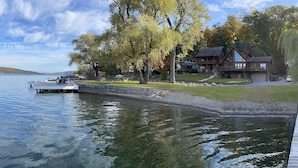 Una propiedad frente al lago con un muelle, una casa grande con terraza, y un área verde con árboles.
