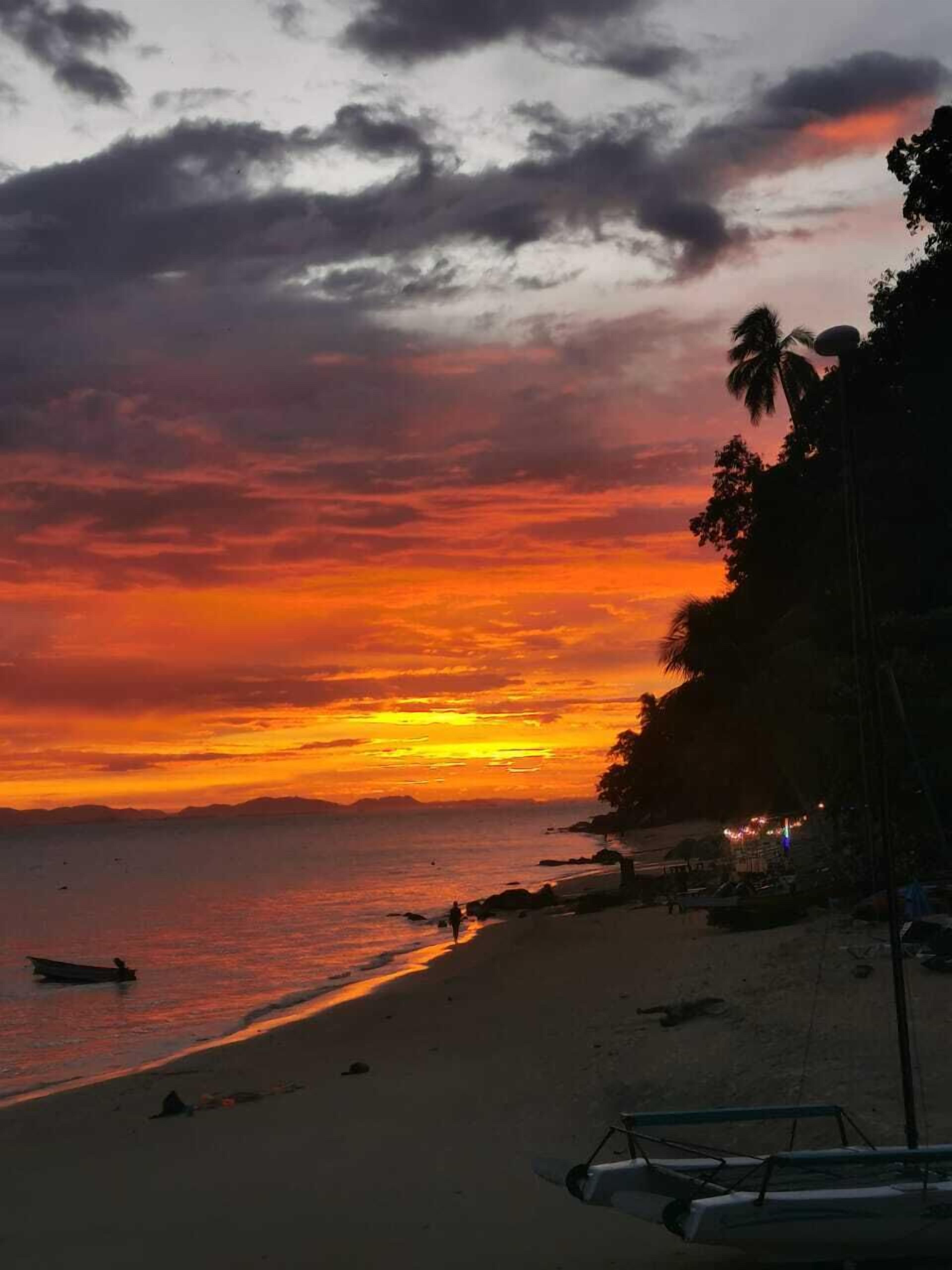 Una playa al atardecer con cielo despejado, mar tranquilo y algunas embarcaciones.
