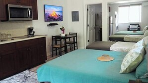 Habitación de hotel pequeña con kitchenette, una cama con colcha color esmeralda, una pequeña mesa de comedor y un televisor montado en la pared.
