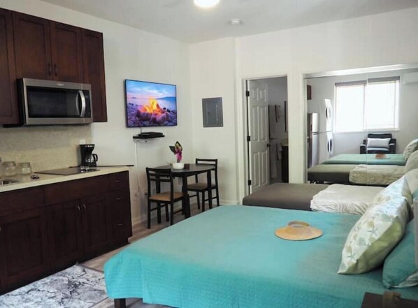 Une chambre d’hôtel compacte comprenant un coin cuisine, un lit avec une couvre-lit turquoise, une petite table à manger et un téléviseur fixé au mur.