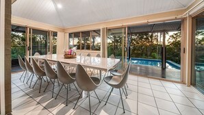 Un espace repas avec une grande table et des chaises, une piscine en arrière-plan et des portes-fenêtres coulissantes donnant sur une terrasse.
