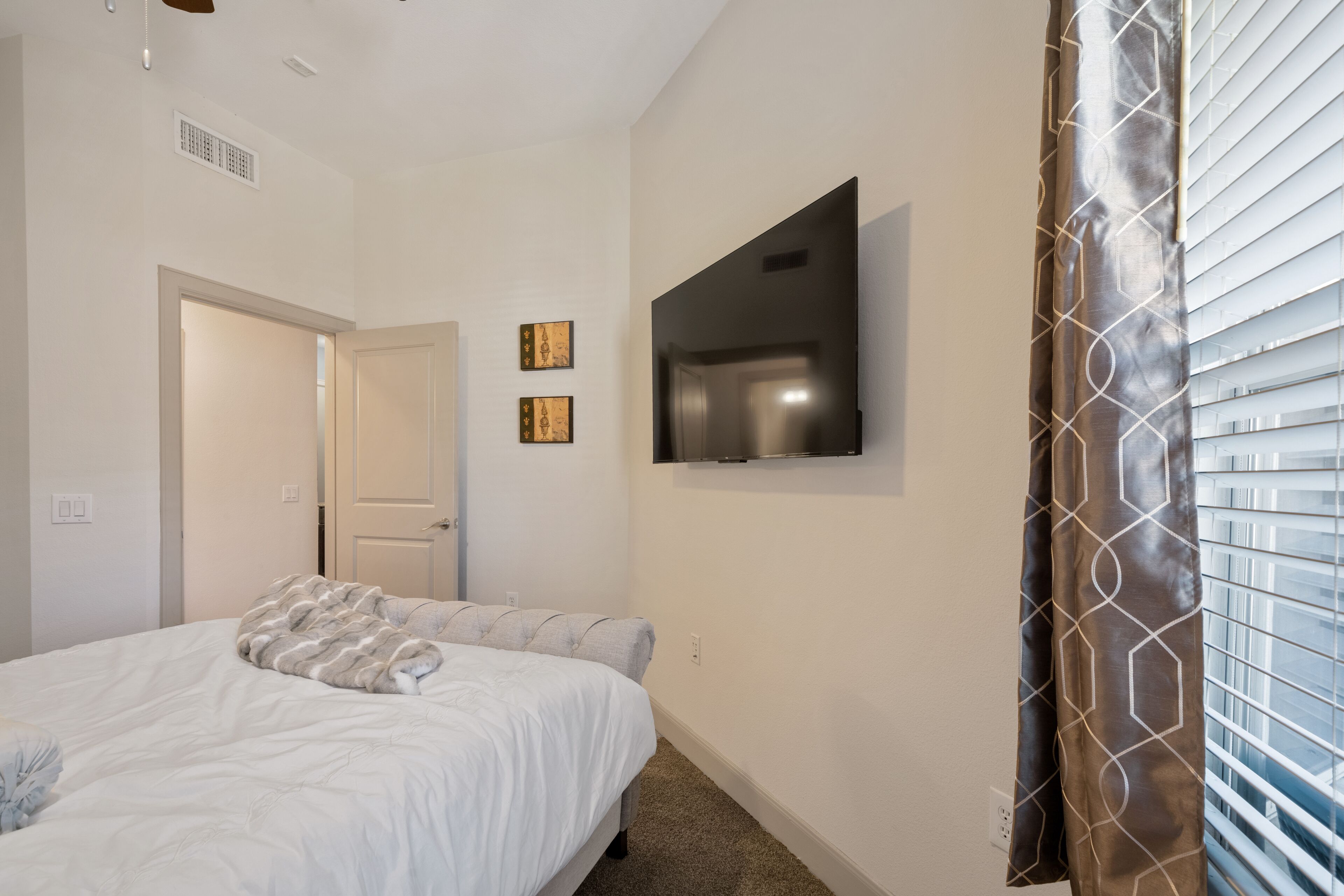 A bedroom with a bed, a TV mounted on the wall, and a window with blinds.