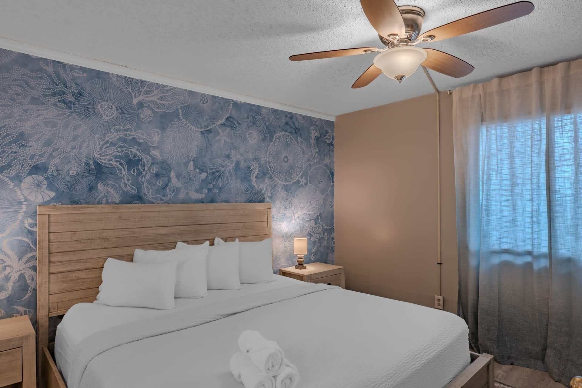 A bedroom with a wooden headboard, a bed with white bedding, a nightstand with a lamp, and a ceiling fan.