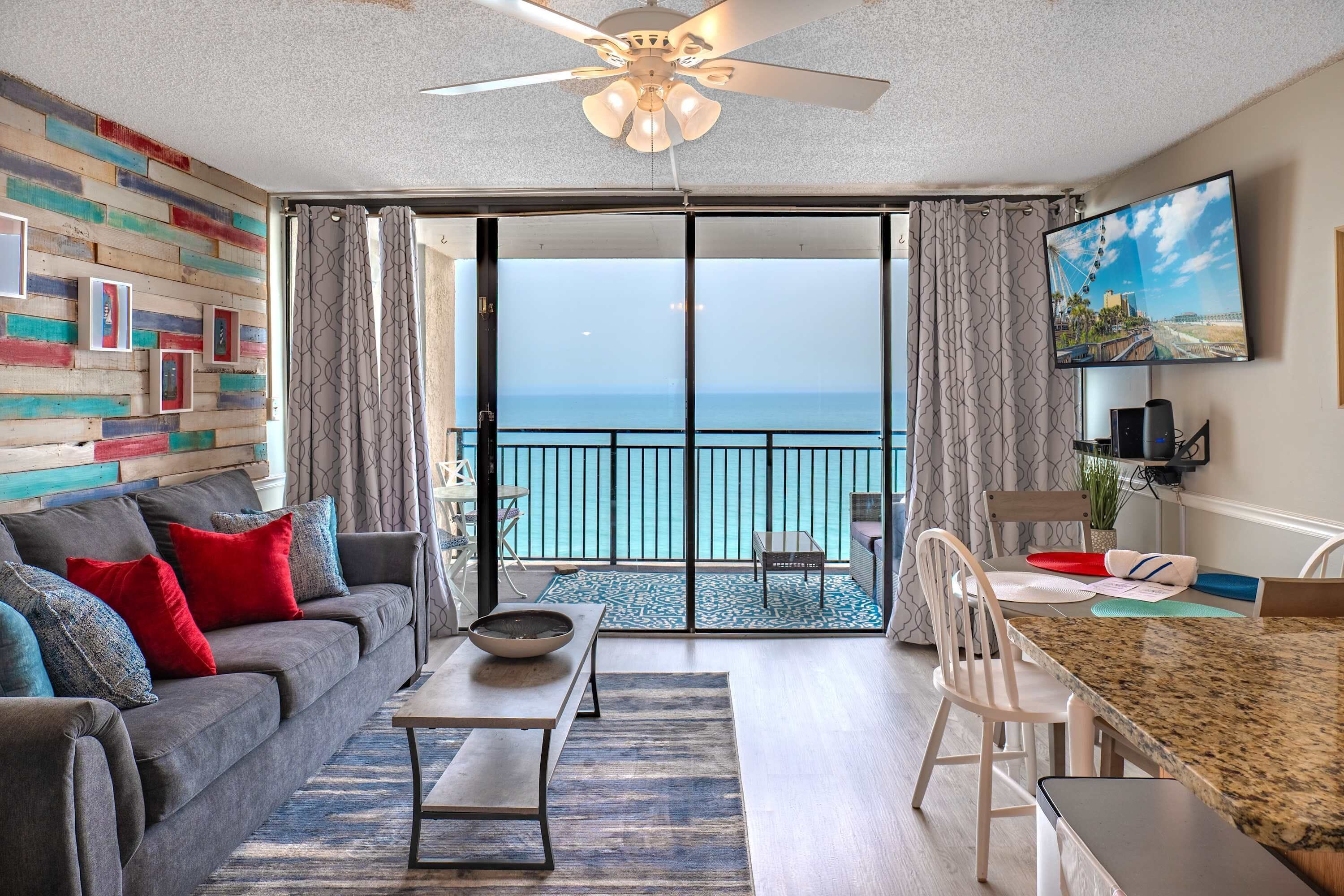A living room with a sofa, coffee table, and dining area with chairs. A large TV is mounted on the wall, and a balcony with a view of the ocean is visible.