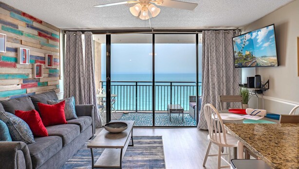 A living room with a sofa, coffee table, and dining area with chairs. A large TV is mounted on the wall, and a balcony with a view of the ocean is visible. - Ocean Forest Plaza 1911 (Myrtle Beach)