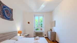 A bedroom with a large bed, a small table, a radiator, and a window with a view of greenery.