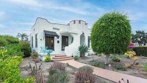 A white building with a curved facade, a small porch with a blue chair, and a well-maintained garden with various plants and a large tree. - Bright Spanish Home in Prime LA Location (Los Angeles)