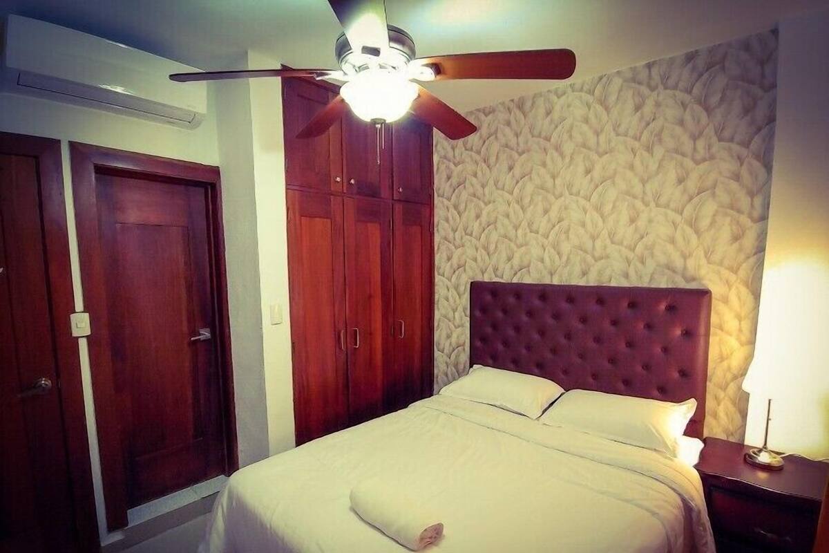 A bedroom with a bed, wooden wardrobe, ceiling fan, and patterned wallpaper.