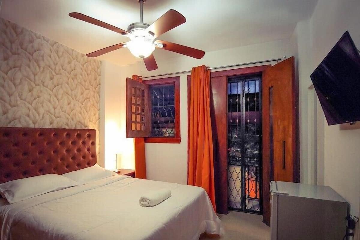 A bedroom with a tufted headboard, a ceiling fan, a bed with a white bedspread, a window with orange curtains, and a flat-screen TV mounted on the wall.