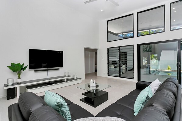 A modern living room with a flat-screen TV, black leather sofas, a glass coffee table, and a large window offering a view of the outdoors.