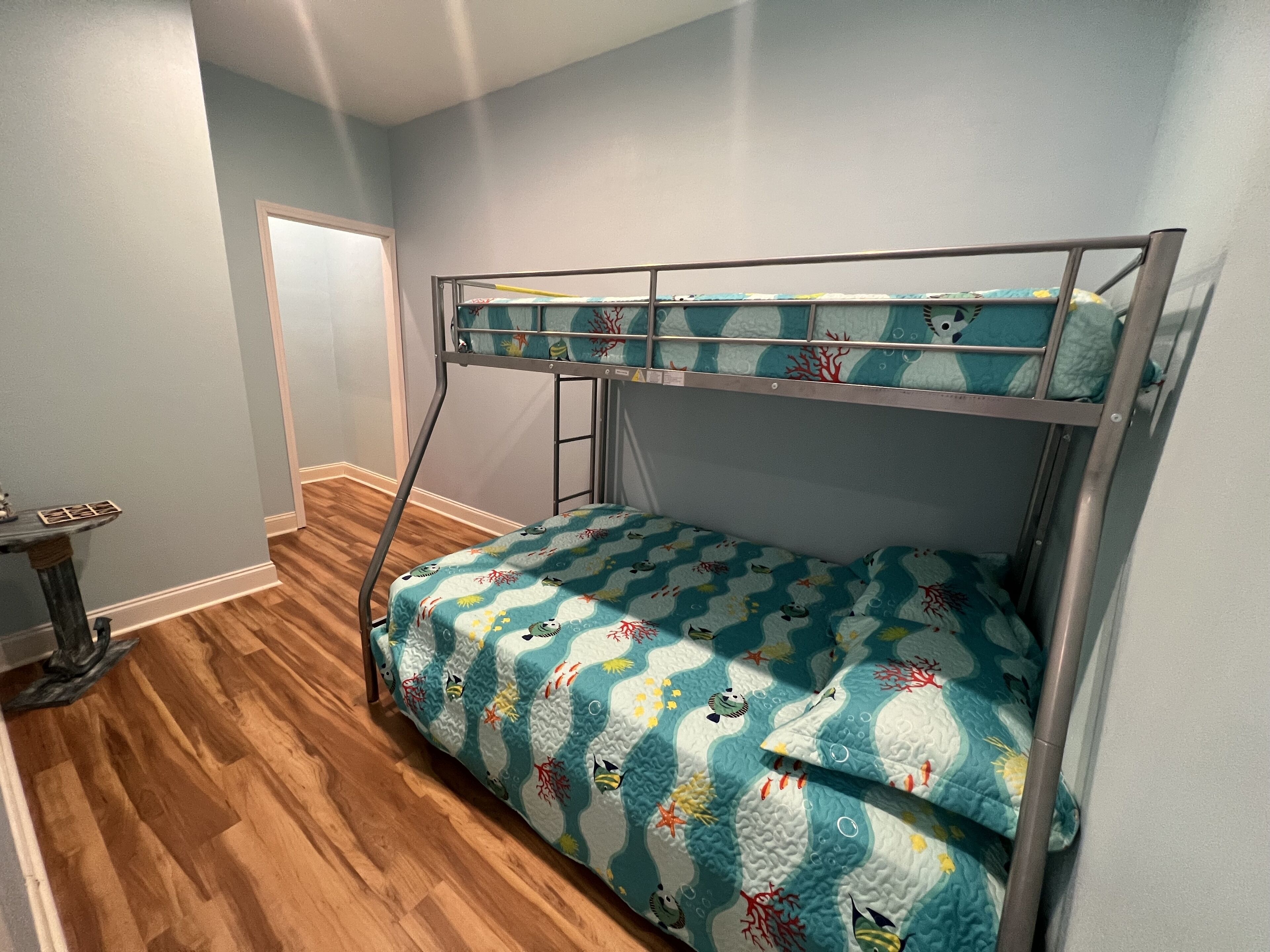 Un lit superposé avec une couvre-lit à motifs bleus et blancs, une petite table avec un pied en métal et un plancher en bois.