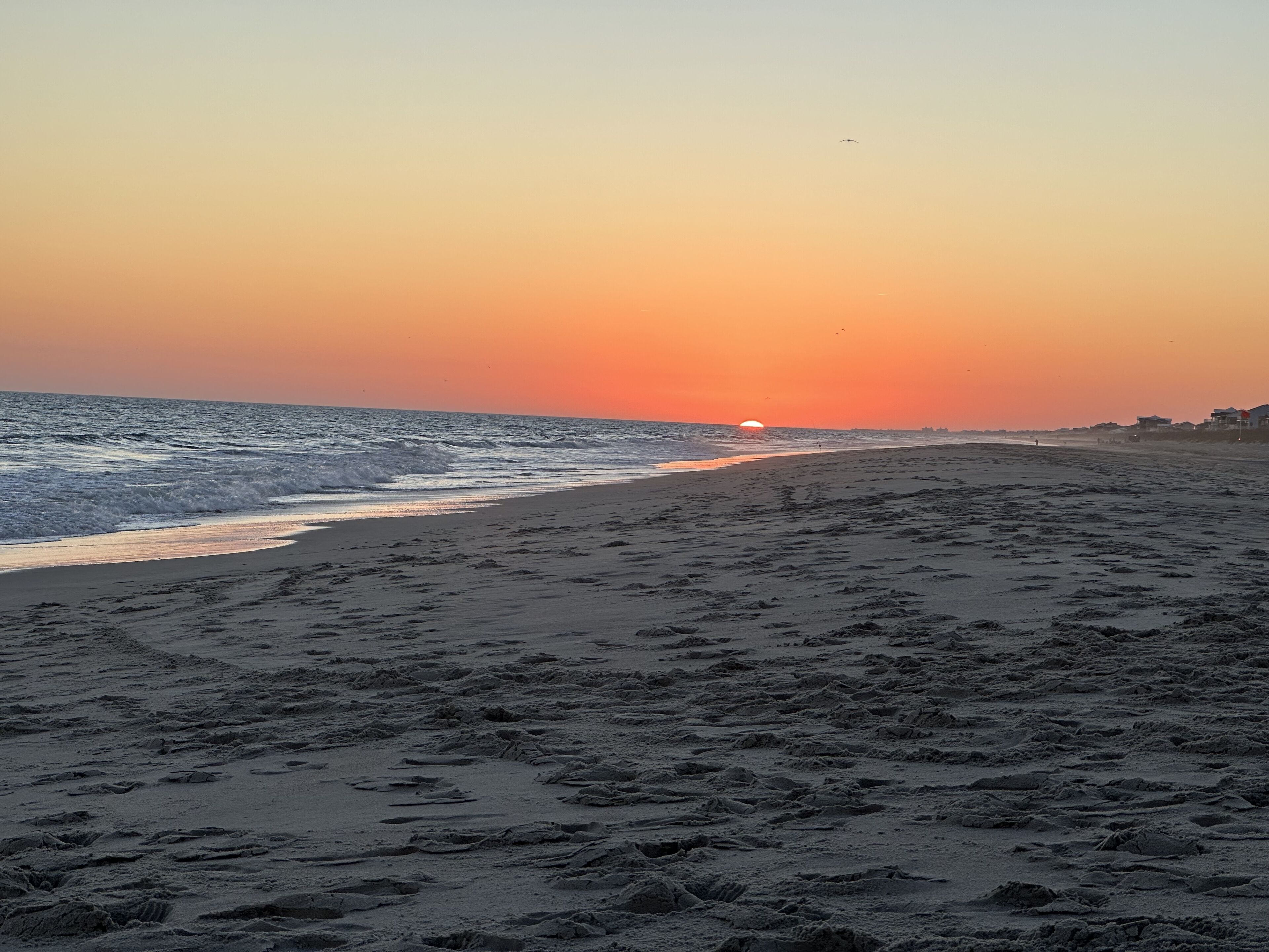 Ein Strand mit Fußspuren, die zum Wasser führen, ein Sonnenuntergang im Hintergrund und wenige Gebäude in der Ferne.