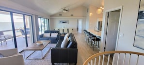 A spacious living area with a large window overlooking a body of water, a black leather sofa, a wooden coffee table, and a bar area with stools.