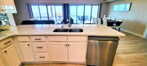 A modern kitchen with a large island, stainless steel dishwasher, and a view of the ocean through large windows.
