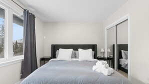 Una cama bien hecha con una colcha a rayas, un cabecero oscuro y almohadas blancas. Se observa un mesita de noche con una lámpara y un espejo, y una ventana con cortina.