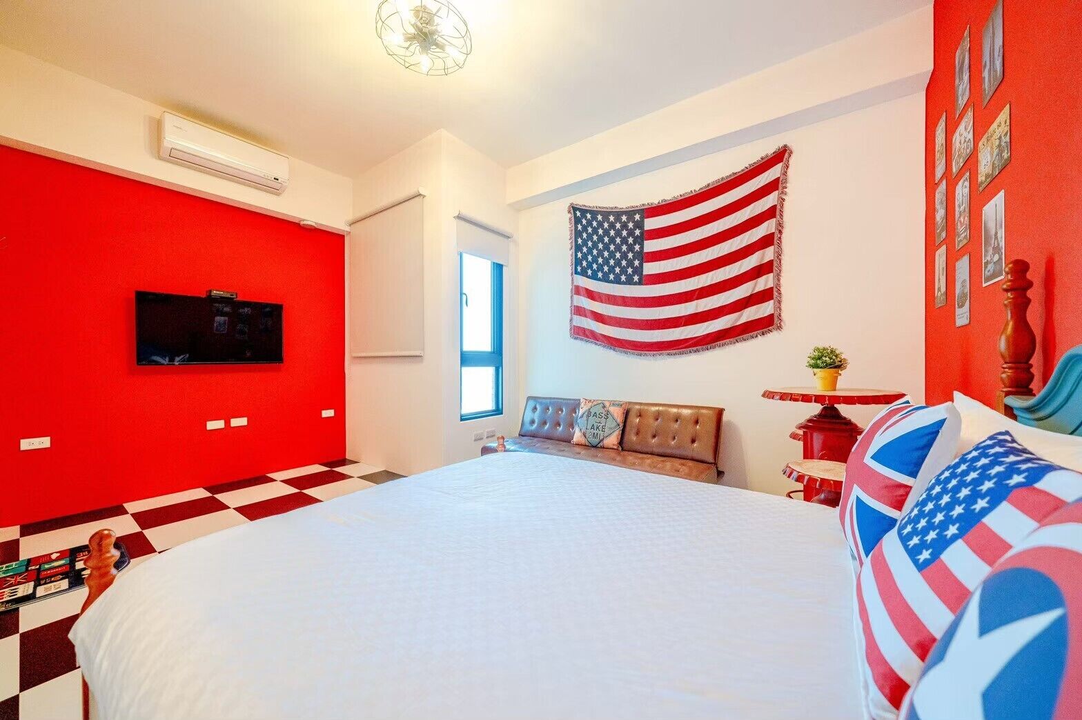 Un dormitorio con una cama grande, una pared de acento roja, una bandera estadounidense, un televisor y un suelo a cuadros.