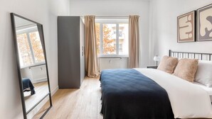 Ein Schlafzimmer mit Bett, Spiegel, Kleiderschrank, einem Fenster mit Vorhängen und Bildern an der Wand.
