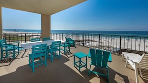 A balcony with turquoise chairs and a table, overlooking a beach with a clear view of the ocean.