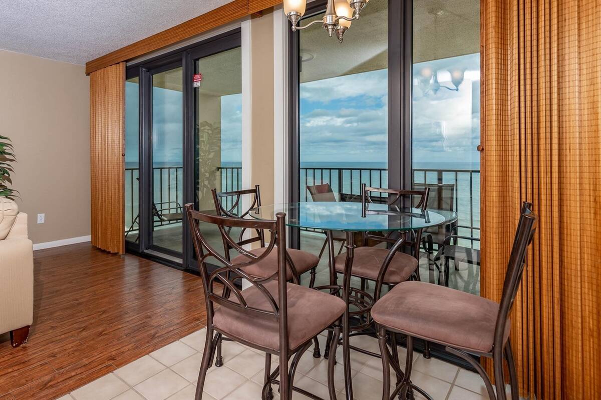 Un coin repas avec une table et des chaises en verre, un lustre et une vue sur l’océan grâce à de grandes portes-fenêtres coulissantes.