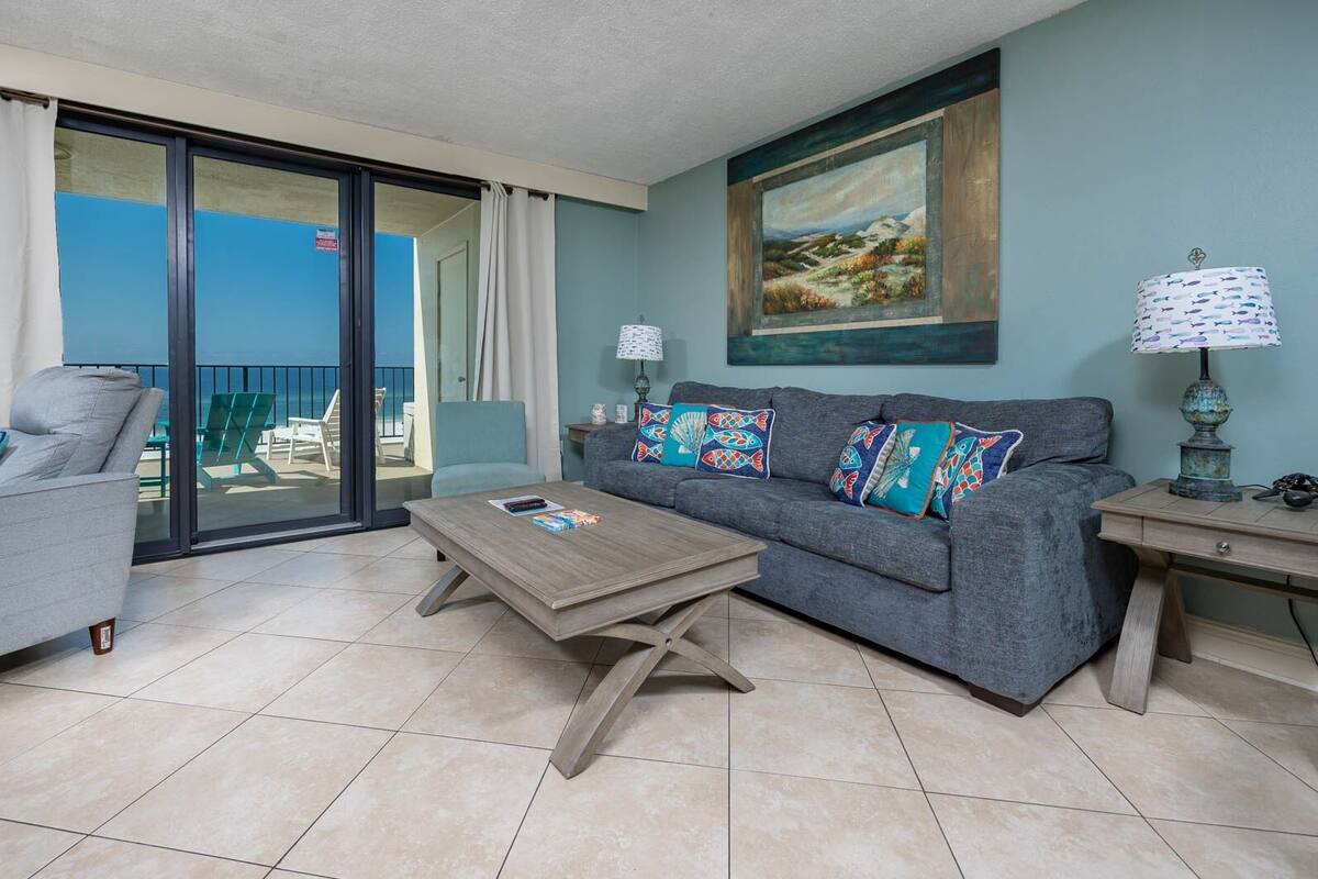 Un salon avec un canapé gris, une table basse en bois et une vue sur la mer, visible à travers une porte-fenêtre coulissante.