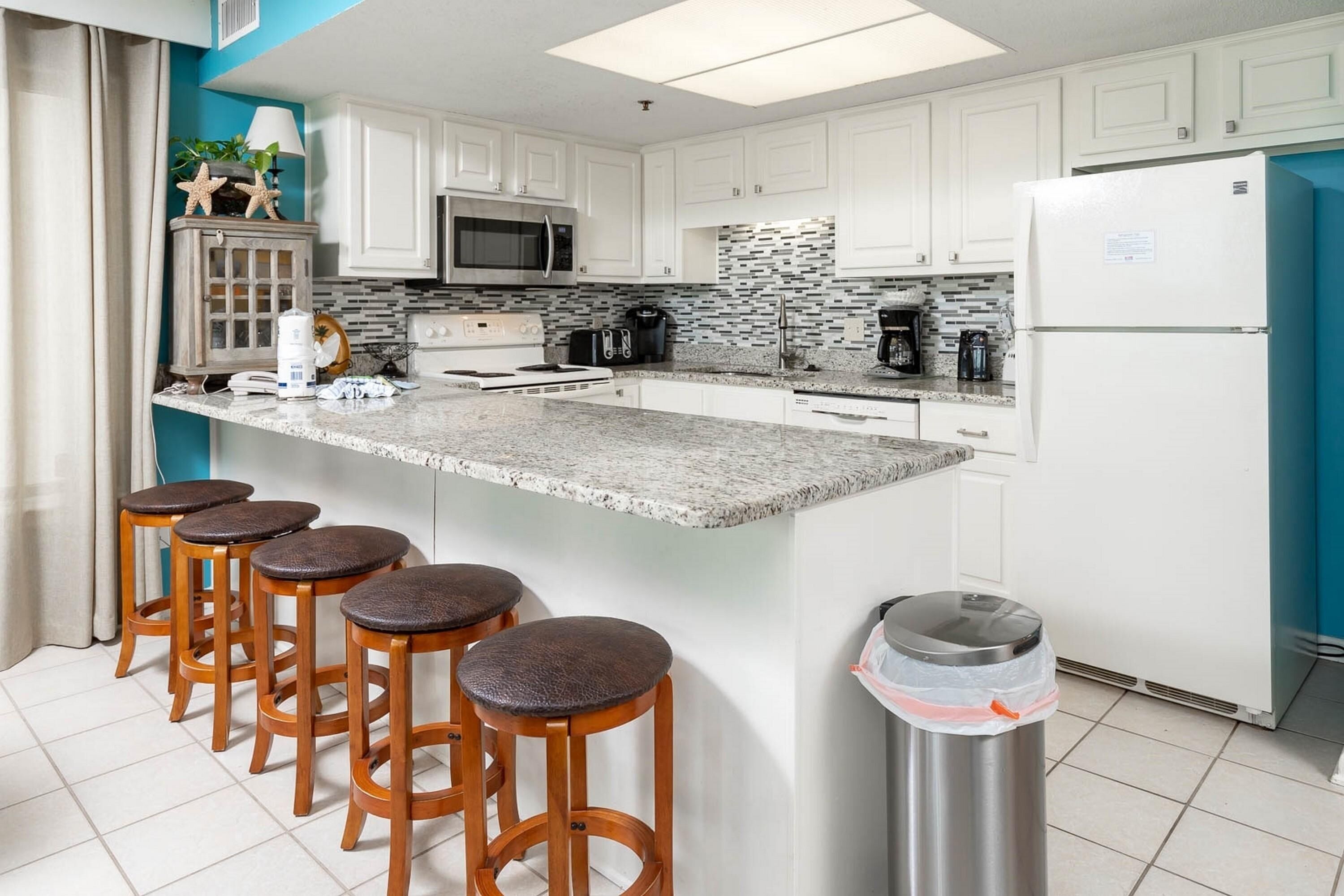A modern kitchen with white cabinets, a granite countertop, a stainless steel refrigerator, and wooden bar stools.