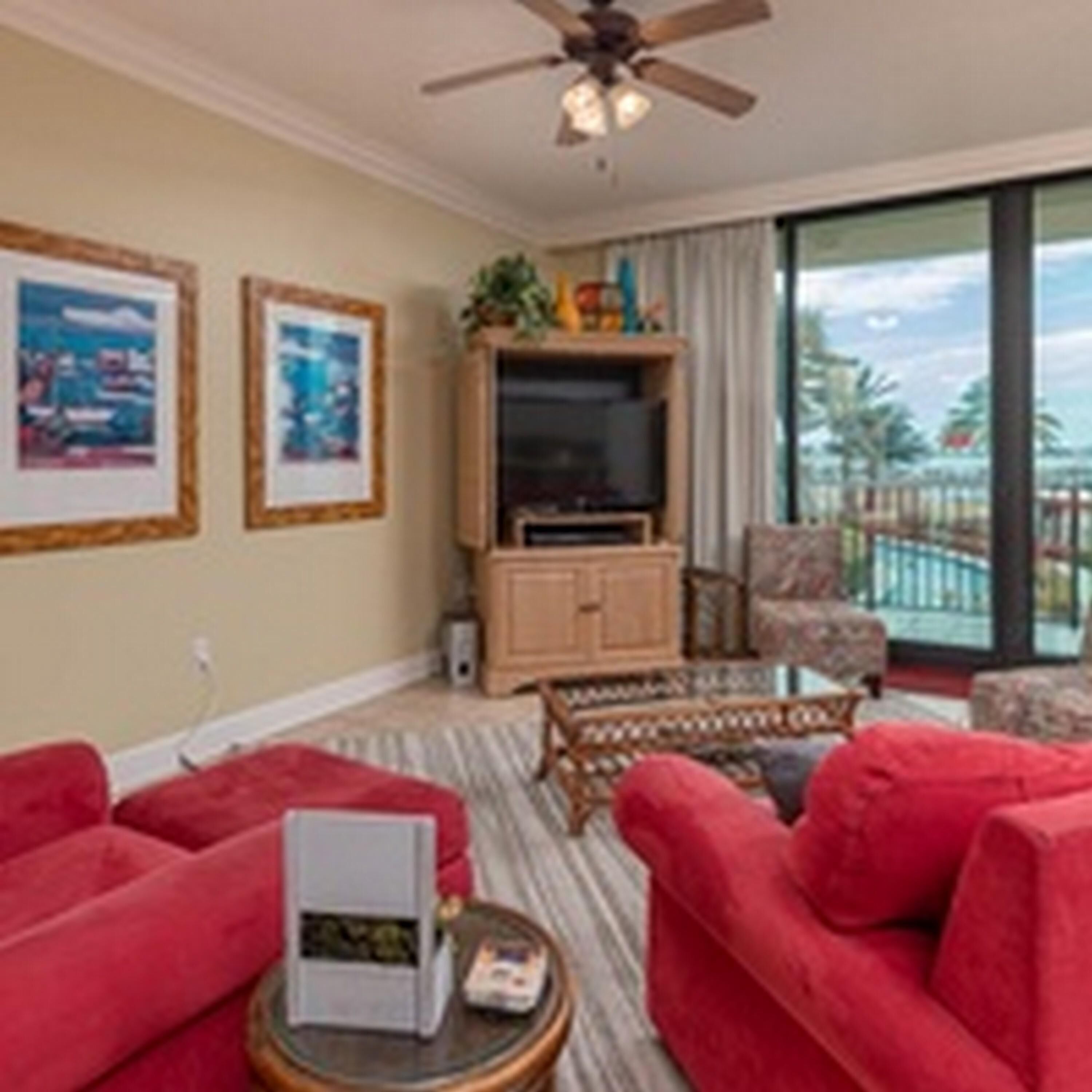 A living room with red sofas, a television, and a balcony with a view of palm trees.