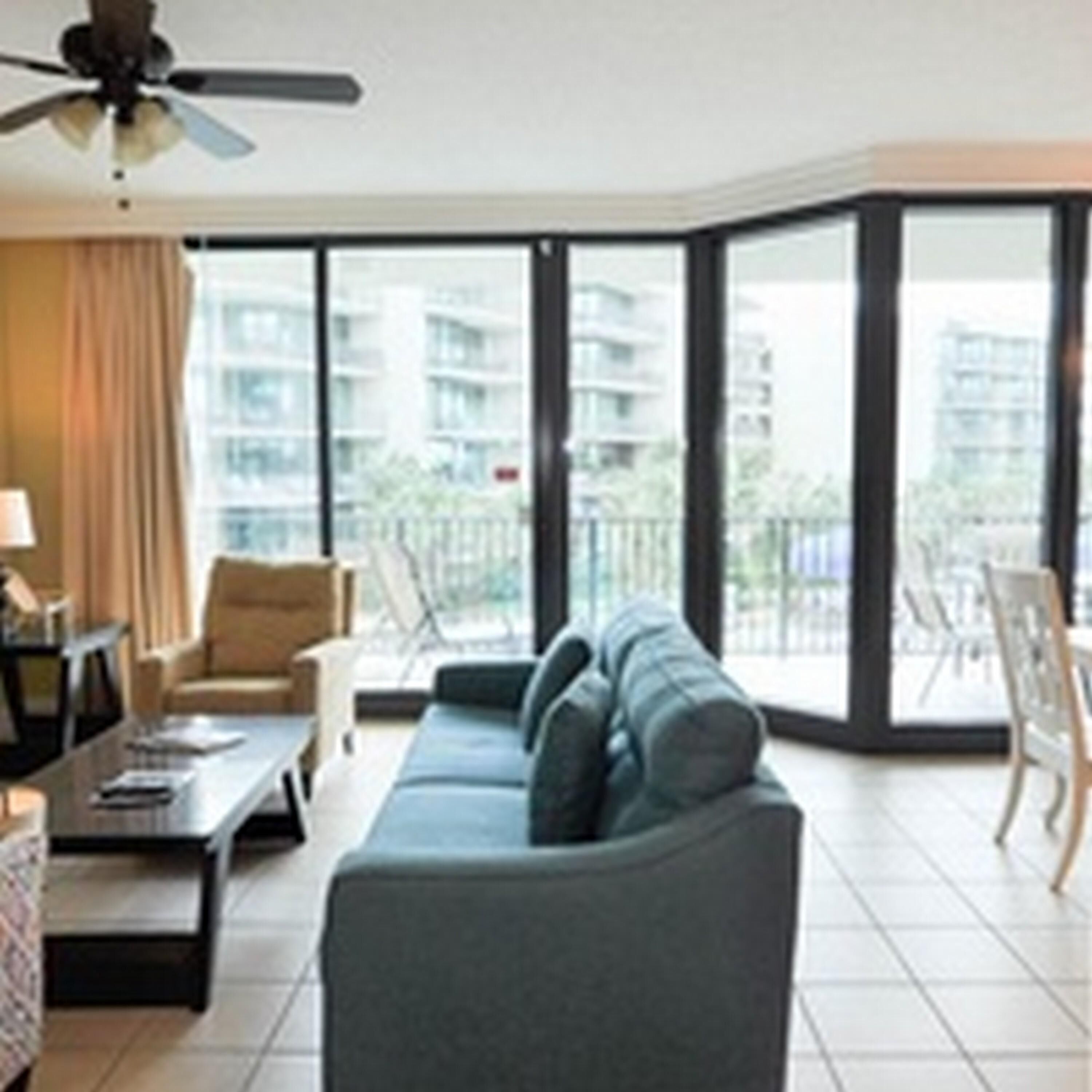 A modern living room with a sofa, coffee table, and chairs. There is a ceiling fan and large windows with a view of buildings outside.