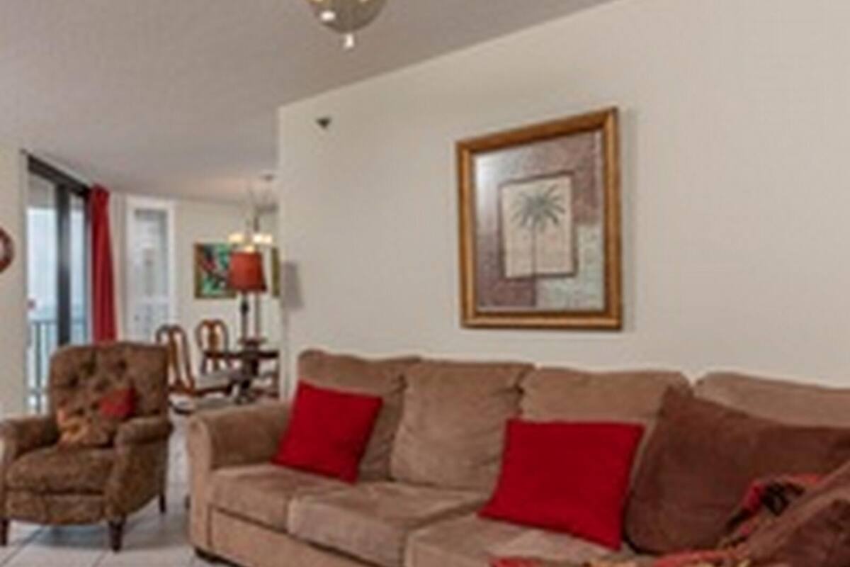 Un salón con un sofá marrón, cojines rojos, un sillón de madera y un cuadro enmarcado en la pared.