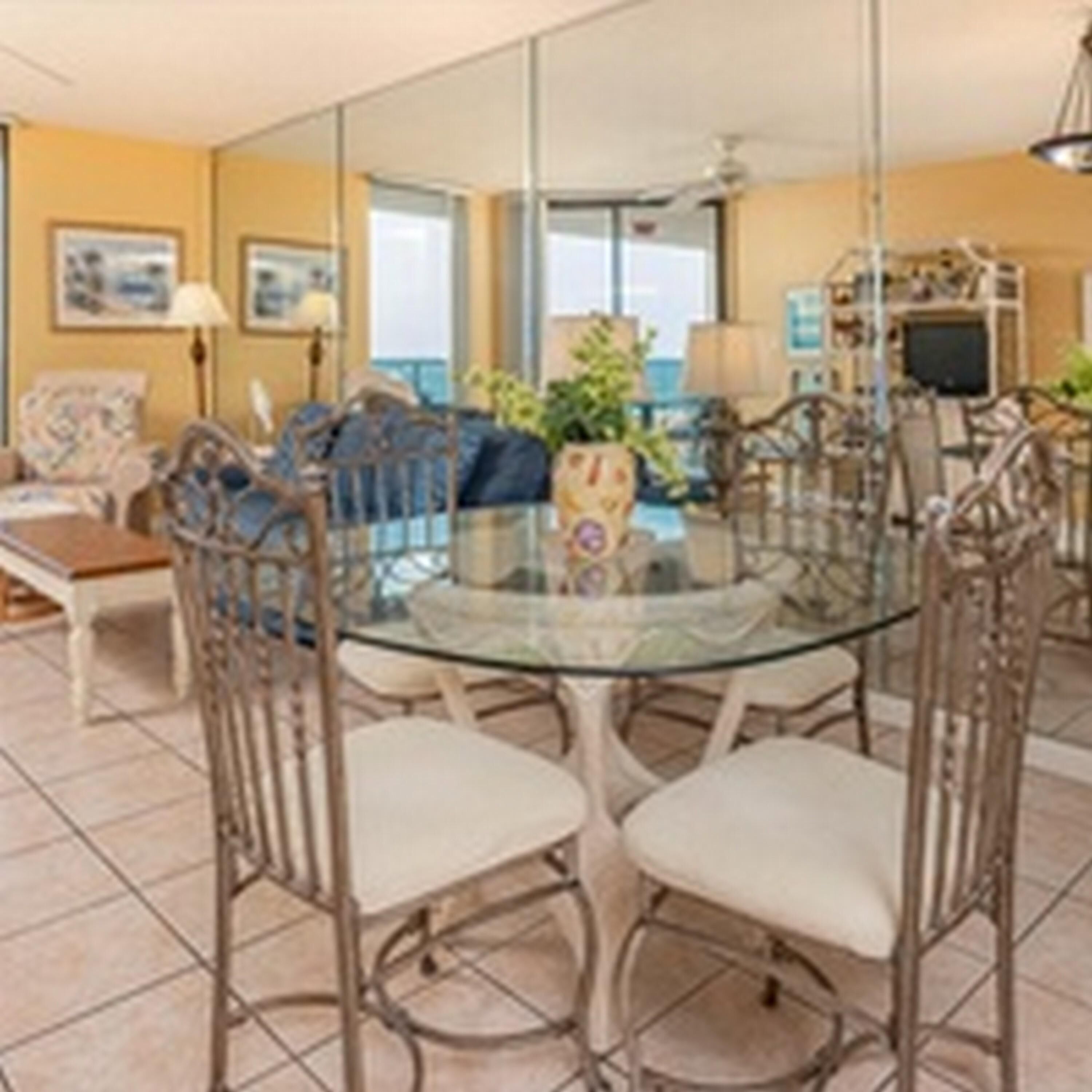 A dining area with a glass table surrounded by chairs, a sofa, and a television in the background.
